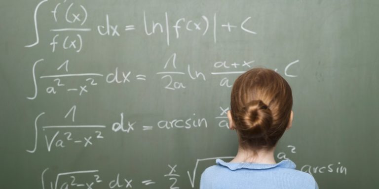 Girl (6-7) standing in front of blackboard, solving arithmetic problem, rear view